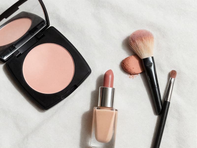 A flat lay of a peach powder palette, brushes and a nude lipstick on linen.